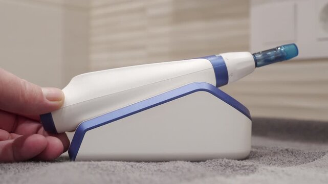 The plastic facial massager is picked up from its charging stand. The device vibrates lightly, emitting a low, continuous buzzing or humming sound.