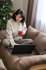 Smiling woman in cozy sweater sits cross-legged on sofa using laptop and holding red mug with decorated Christmas tree in background. Holiday shopping, working from home, cozy winter concept