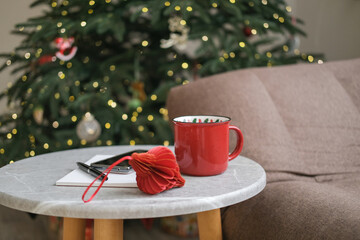 Red mug, notebook with pen and red paper Christmas tree decoration on gray table next to sofa with blurred Christmas tree in background. Holiday break, cozy winter, planning, festive atmosphere