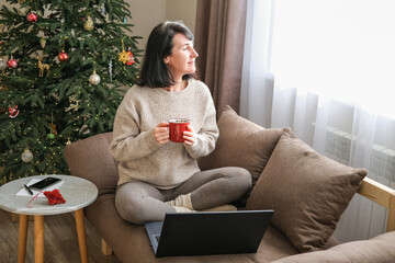 Woman in sweater sits cross-legged on sofa with laptop, holding red mug and looking out sunlit window with decorated Christmas tree in background. Relaxation, winter holidays, cozy home life