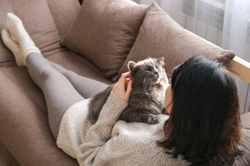 Woman is lying on brown sofa cuddling and stroking fluffy gray cat resting on her chest. Pet companionship, relaxation, cozy home life and comfort with animals