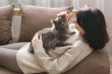 Woman is lying on sofa, gently petting fluffy gray cat resting contentedly on her chest. Pet companionship, relaxation, comfort, human-animal bond