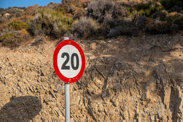 Speed limit sign near rocky hillside within bright sunlight, featuring dry shrubs above