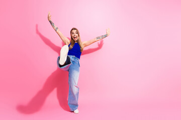 Young woman with tattoos wearing blue top and jeans jumping against pink background smiling and celebrating fashion lifestyle