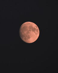 A close-up of the Moon in the waning or growing phase, illuminated by a warm orange-red light that highlights the details of the lunar seas and craters against the background of the deep night sky.