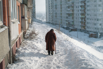 Fototapeta premium An elderly woman in a coat walks through the snow leaning on a cane in a winter city