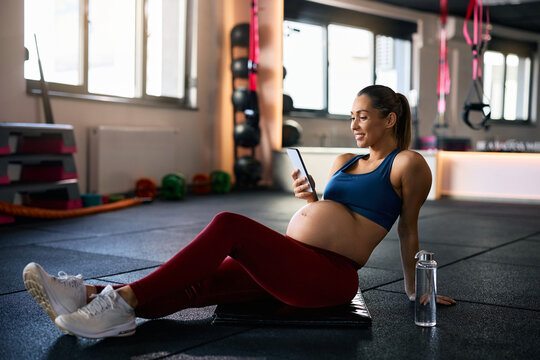 Happy pregnant athlete using cell phone during her water break at fitness studio.