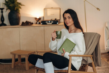 Beautiful brunette is with cup of tea. Relaxed woman is sitting in the chair and reading the book at home