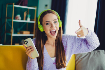 Happy young woman with green headphones on a phone giving a thumbs up at home in a cozy living room in a bright modern setting