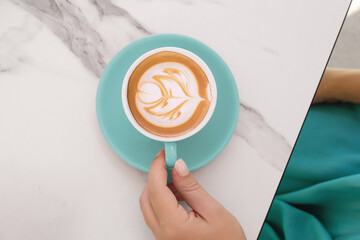 A hand holding a turquoise coffee cup with elegant latte art on a marble table.