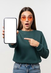Surprised woman with orange glasses pointing to a blank smartphone screen for advertising or app promotion in a studio shot against a neutral background