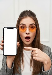 Surprised woman with orange glasses pointing to a blank smartphone screen for advertising and marketing in a modern and clean studio setting
