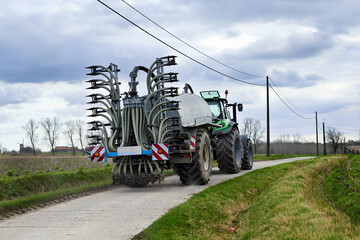 A green tractor pulls a large farm implement on a rural road under a cloudy sky. Power lines...