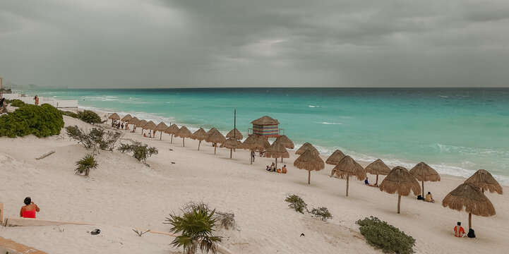 Wide sandy beach with turquoise waves and dramatic cloudy sky. Tourists walking along the shore, tropical coastline and moody weather atmosphere.