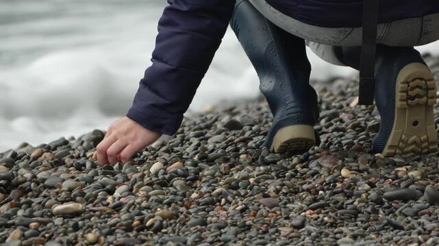 Boots, pebbles, beach. Woman in rubber boots carefully picking up smooth stones near the crashing sea.