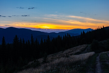 Obraz premium A serene twilight panorama of layered mountains and dark conifer silhouettes beneath a vivid orange and blue sunset, with a dirt path across a grassy hillside inviting wonder and calm reflection.