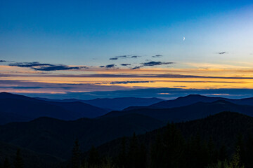 Blue hour over layered mountain ridges with a crescent moon and glowing horizon. Deep tones and serene clouds create a peaceful, contemplative wilderness vista perfect for travel and backgrounds.