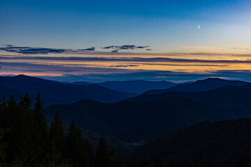 Layered mountain ridges fade into twilight under a delicate crescent moon, with dark spruce forest below and a glowing horizon of blue and amber clouds, creating a serene panoramic wilderness now