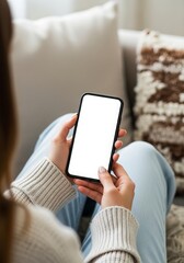 Woman holding a blank smartphone with a white screen while relaxing on a couch in a cozy living room setting perfect for showcasing mobile app designs