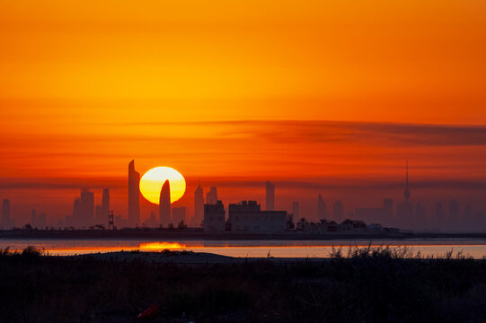 The sun is setting over a Kuwait city skyline, casting a warm glow over the buildings.
