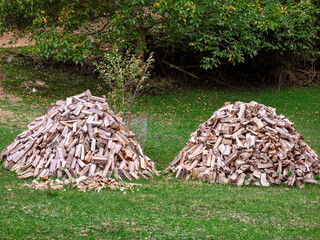 Two large piles of chopped firewood stacked on green grass in a rural backyard. Dry split logs prepared for winter heating. Natural rustic scenery with trees and autumn leaves in the background.