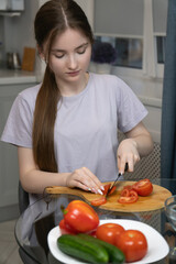 Focused young girl neatly chopping fresh tomatoes on a wooden cutting board at a glass table, preparing a healthy meal or salad with various vegetables in her home kitchen