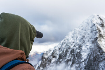 man hiker wearing a green hoodie and cap, gazes at a snow-covered mountain range under a cloudy sky, winter exploration