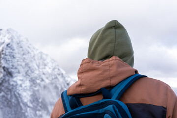 person with a green hood and brown jacket stands against a snowy mountain backdrop. The scene captures a sense of adventure in a winter landscape, hiking