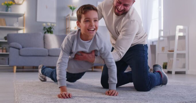 Smiling father helping young son to practice push ups on carpet in living room. Caring parent supporting preteen boy in developing strength, encouraging fitness and healthy lifestyle at home workout.