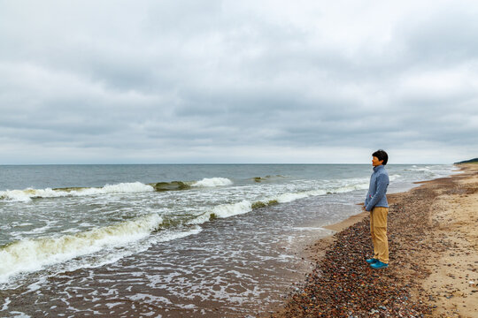 Person standing on a windy and rugged beach looking at the wavy ocean under a dramatic, overcast gray sky, with a mix of sand and pebbles