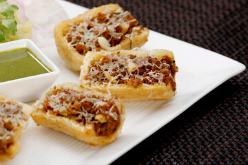 Close-up of golden-brown Indian savory snacks (Khasta Kachori) made from puff pastry with a spiced filling, topped with fine garnish, served next to a small bowl of vibrant green chutney.