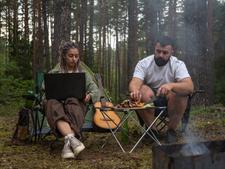 Freelancers enjoying camping trip in nature, combining work and leisure, with man preparing barbecue and woman using laptop near tent and guitar