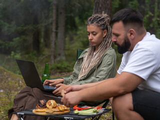 Young woman working on laptop while camping in forest, while man preparing barbecue food, enjoying the outdoor lifestyle and remote work opportunities