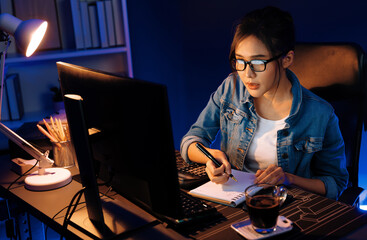 Thinking young beautiful Asian creative woman looking on pc with taking note on paper for memo in sales target report on business market graph report at neon light modern office at night. Stratagem.