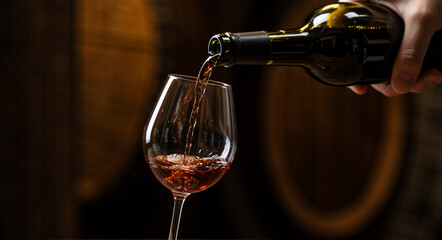 Pouring red wine into a glass with wooden barrels in the background. Winery cellar tasting concept