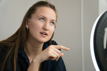 Young woman sitting on the bed, recording a video with her smartphone and ring light, gesturing with her finger while explaining concepts in an engaging manner