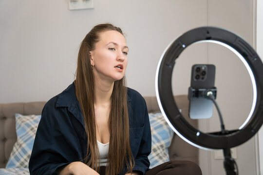 Young woman beauty blogger applying makeup in front of a ring light while recording a video for her followers on social media, sharing tips and techniques for cosmetics enthusiasts
