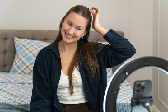 Young woman sitting on her bed, adjusting her hair and preparing for a live stream. Ring lamp illuminating her face as she sets up her smartphone for an engaging broadcast