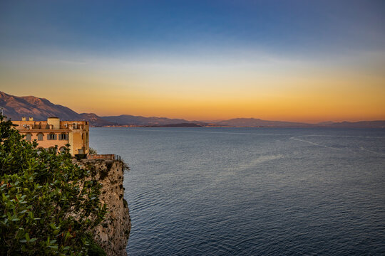 Gaeta, Latina, Lazio, Italy. A beautiful sunset over the sea, seen from a panoramic viewpoint in the village. A building overlooking the sea, perched on a sheer cliff. Mountains in the background. - Powered by Adobe