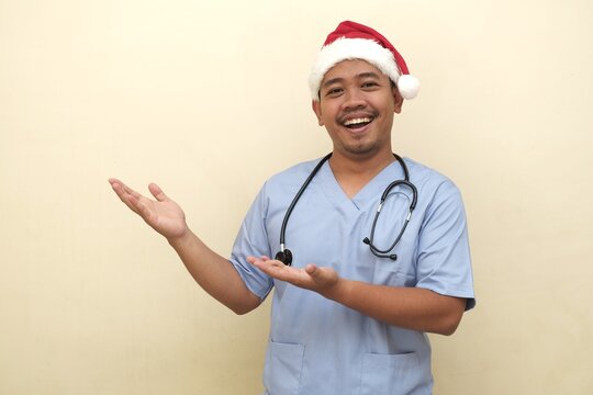 Southeast Asian male nurse in Santa hat and scrubs smiling and gesturing as if presenting something, isolated and ideal for holiday ads.
 - Powered by Adobe
