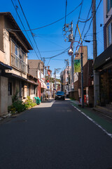 Fototapeta premium A narrow Kamakura street shows wooden and concrete buildings, power lines, a small truck, vending machines, and plants in bright midday sun with crisp shadows.