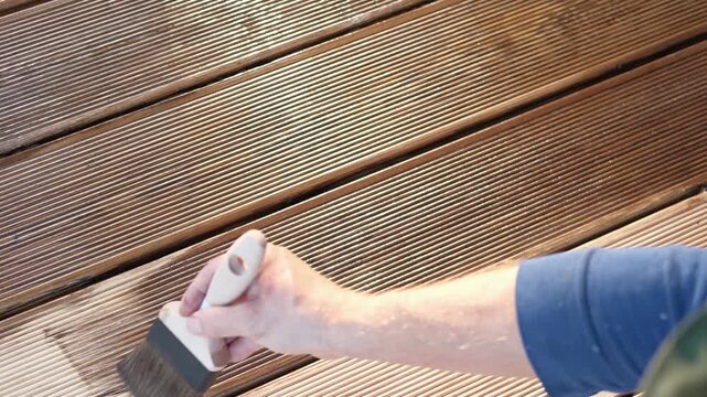 Brown oil for protection of wood by staining. Painting a larch for protecting and pests. Woodworker applying finish to terrace deck board. Repairman varnishing with brush wooden plank floor surface.
