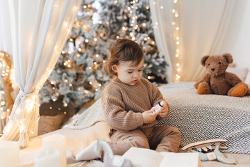 Little boy sitting on the bed in front of christmas tree. Toddler playing with toys.