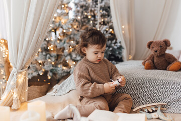 Little boy sitting on the bed in front of christmas tree. Toddler playing with toys.