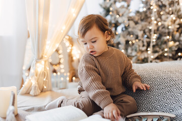 Little boy sitting on the bed in front of christmas tree. Toddler playing with toys and candles. Brown colors.
