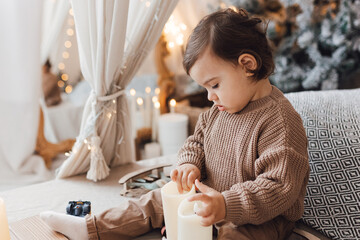 Little boy sitting on the bed in front of christmas tree. Toddler playing with toys and candles. Brown colors.
