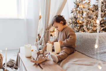 Little boy sitting on the bed in front of christmas tree. Toddler playing with toys and candles. Brown colors.