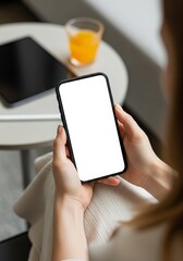 Woman holding a smartphone with a blank white screen sitting near a table with a tablet and orange juice ideal for app promotion and mobile technology display