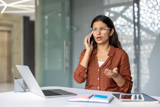Indian businesswoman in eyeglasses having a serious and concerned phone call, discussing problems while working on her laptop in a modern office environment