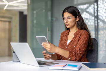 Young businesswoman in glasses concentrates on laptop and tablet, multitasking in a bright modern office to manage projects, plan strategy and boost productivity